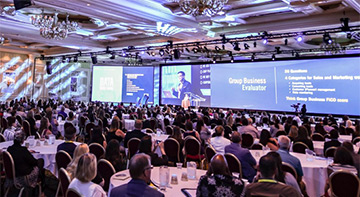 A conference room with a large audience in front of a large screen and a speaker.