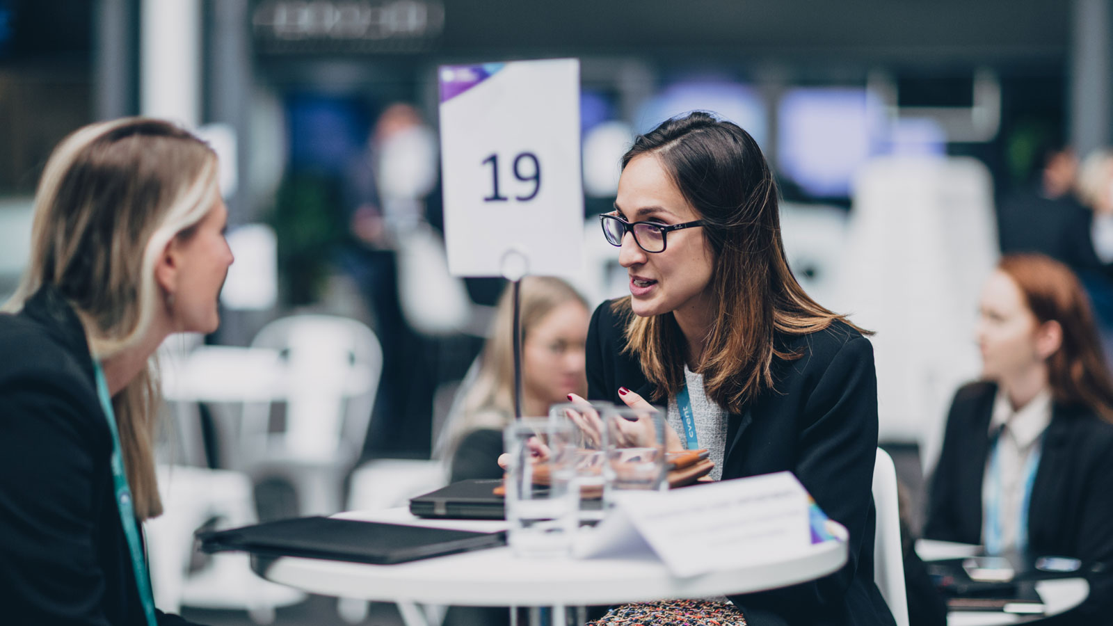 Two women having a conversation at a table with a sign reading 19 behind them.