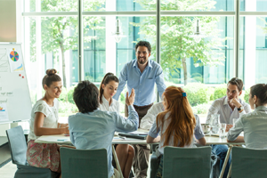 Group of people sitting at a table in a conference room with a large window and trees outside.