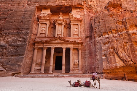 Camels with red saddles and a man standing near the Al Khazneh temple in Petra, Jordan.
