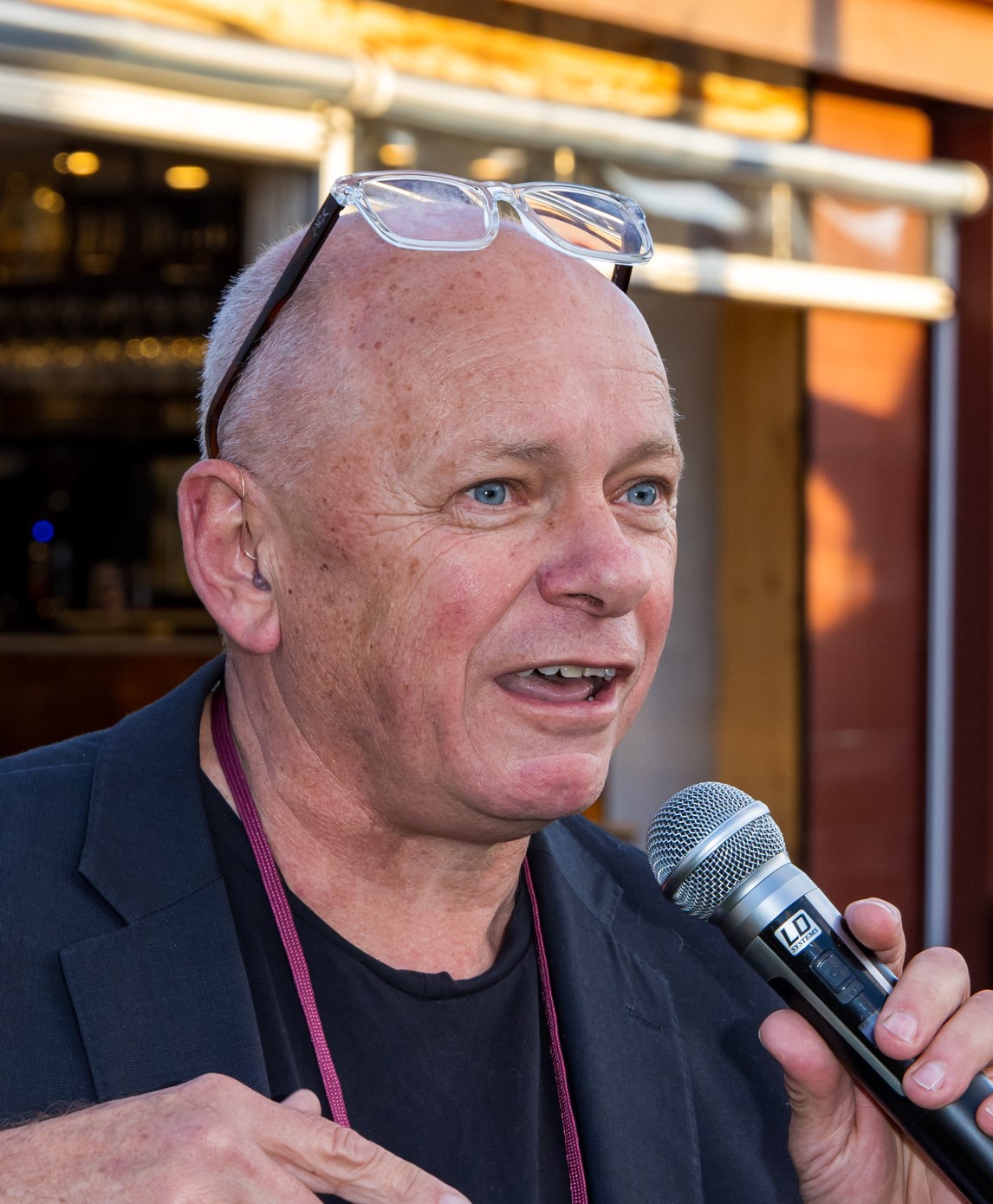 Brett, a man with a microphone, is talking and standing in front of a bar with a yellow light.