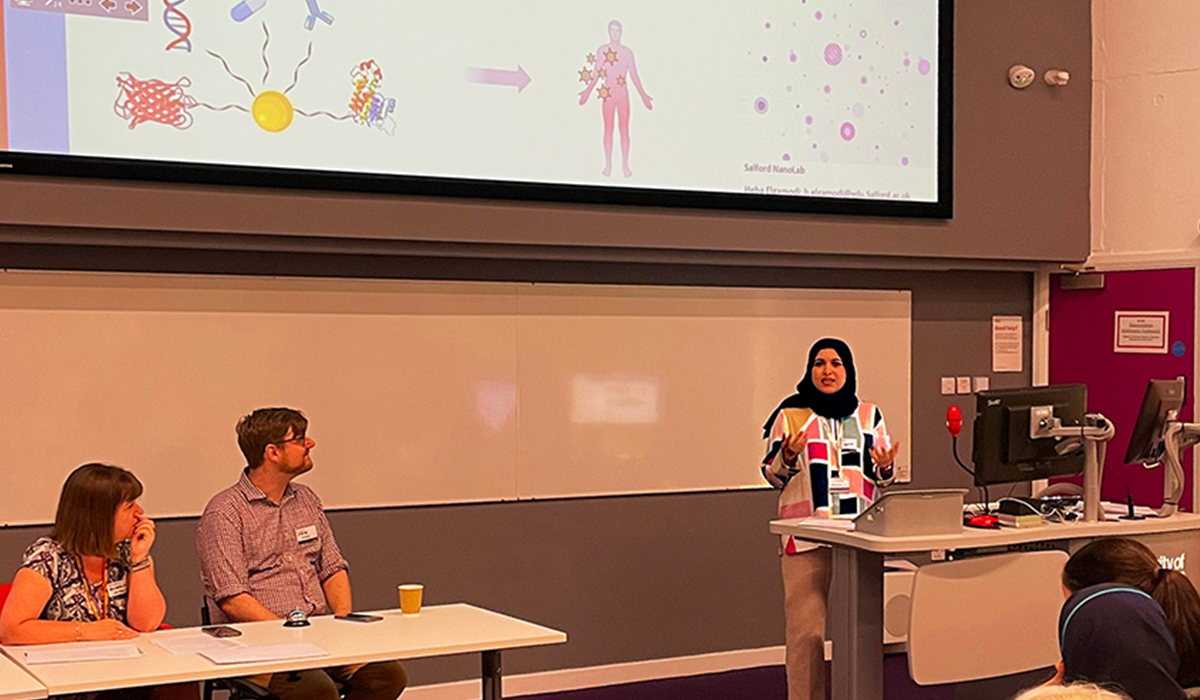 Two people sit at a table, while a woman stands at a podium in front of a screen.