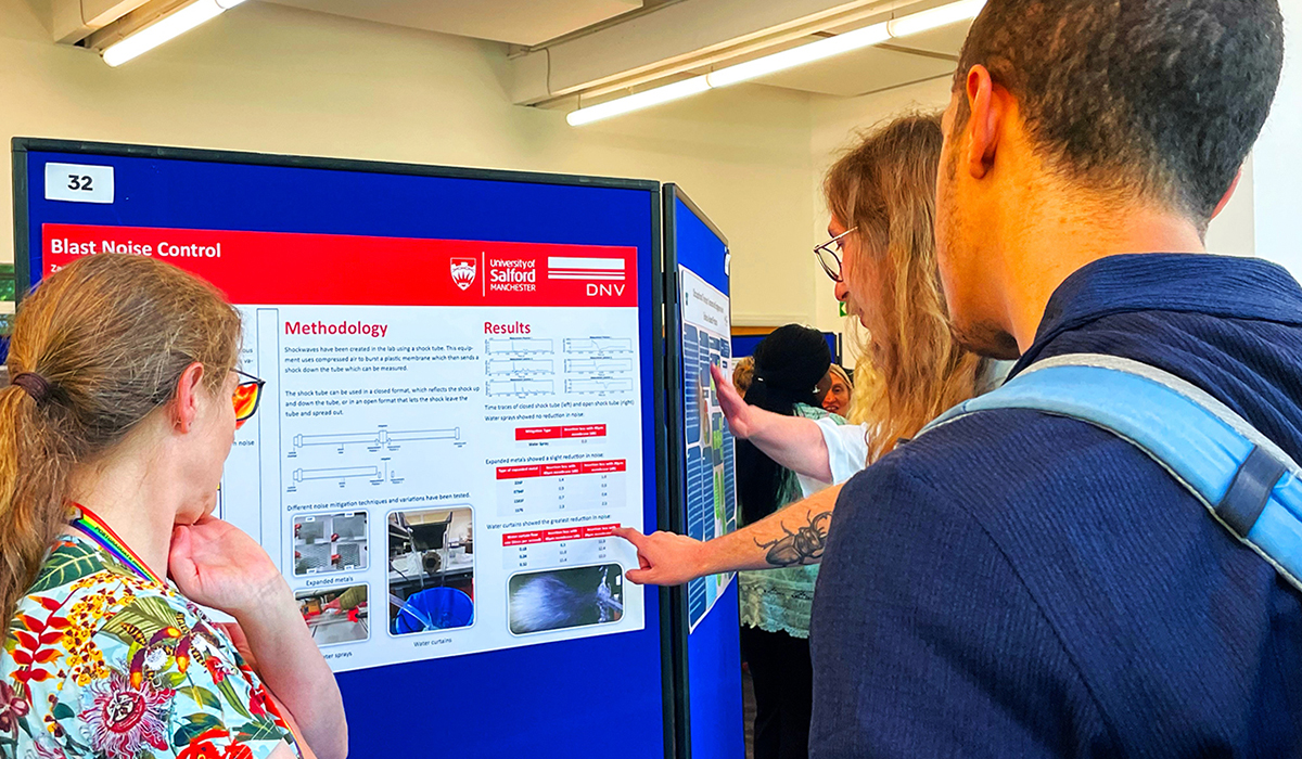 A group of people at an exhibition stand looking at a poster titled 'Blast Noise Control'.
