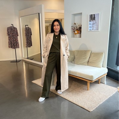 A woman in a trench coat stands in front of a mirror in a room.