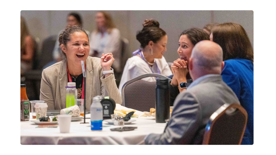 Two people are seated at a round table in a conference or event setting. The person on the right is smiling and wearing a dark shirt with a purple lanyard, while the person on the left is listening attentively with their hand resting on their chin. The background is softly blurred, showing other attendees and tables, with purple and pink lighting creating a vibrant atmosphere. Items such as cups, a phone, and glasses are visible on the table in the foreground.