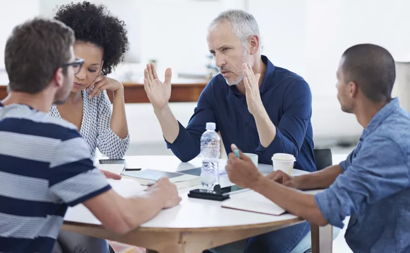 Three people sitting around a table with a man raising his hand in the middle of the table.