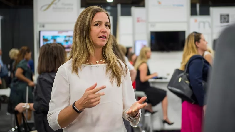 A woman stands in a trade show surrounded by other women, booths, and monitors.