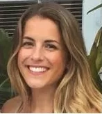 Blonde woman with long hair smiling and posing for a picture in front of a plant.