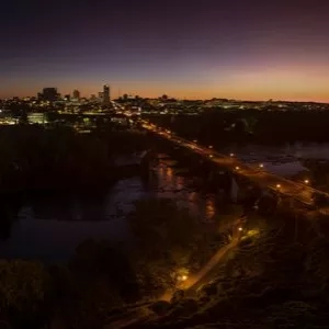 Aerial view of a city at dusk, showing the skyline, a bridge, and a river.