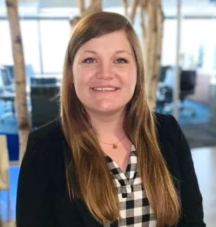 A smiling woman in a black blazer and checkered shirt stands in front of a glass wall.
