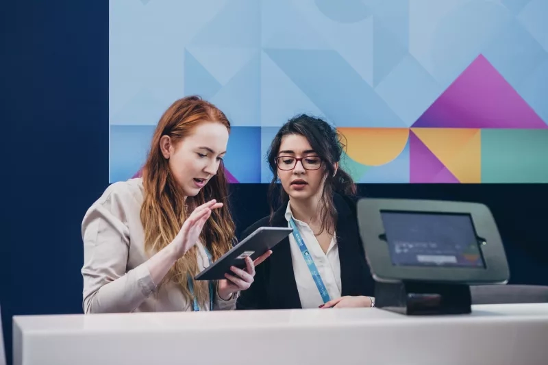 Two women wearing name tags and looking at a tablet in front of a colorful screen.