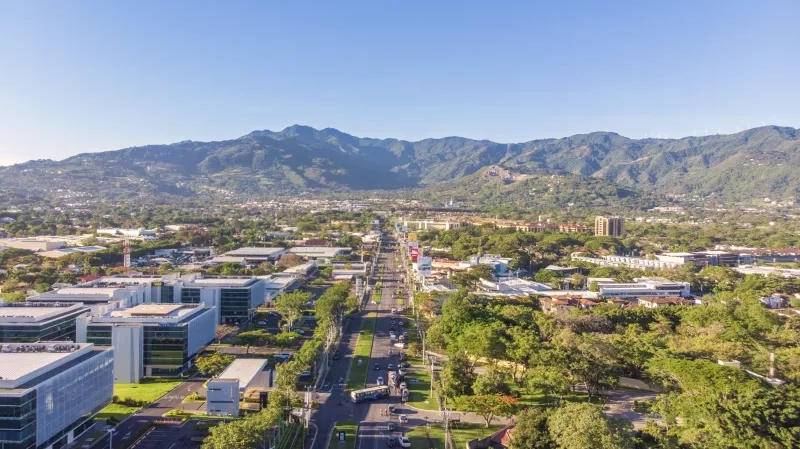 A city view with buildings, mountains, and a wide road with cars and trees.