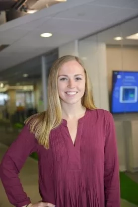 A smiling woman in a purple blouse stands in an office, with a TV in the background.