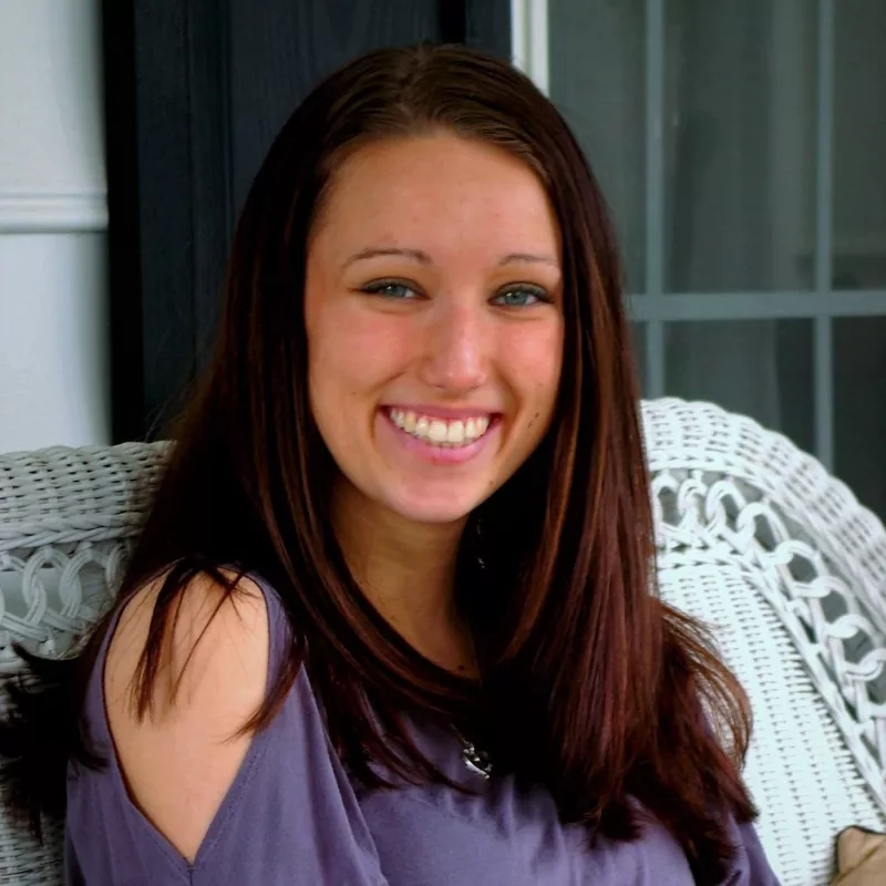 A smiling woman with brown hair and blue eyes sits on a white wicker chair in front of a window.