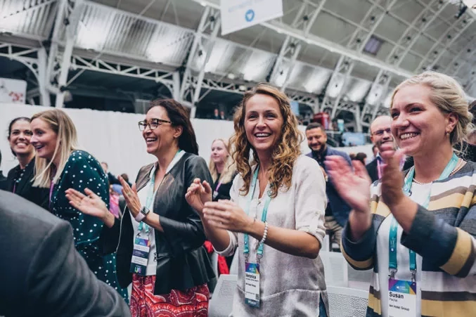 Women at the front of an audience clap, smiling, during a conference at the Tobacco Dock.