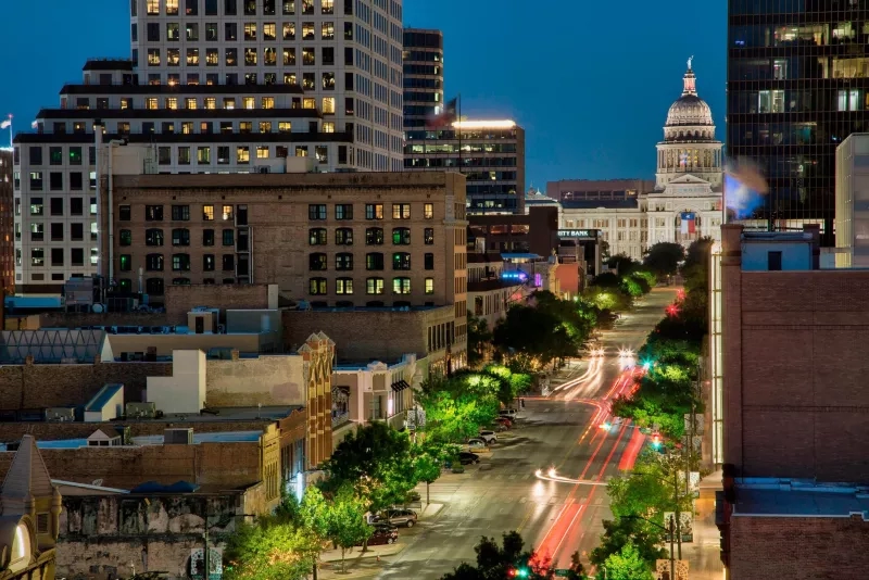 Cityscape at night with tall buildings, a dome-shaped structure, trees, and a street with red lights.