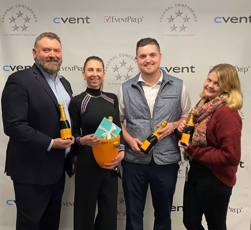 Four people posing for a picture holding bottles of champagne in front of a Federal Conference backdrop.