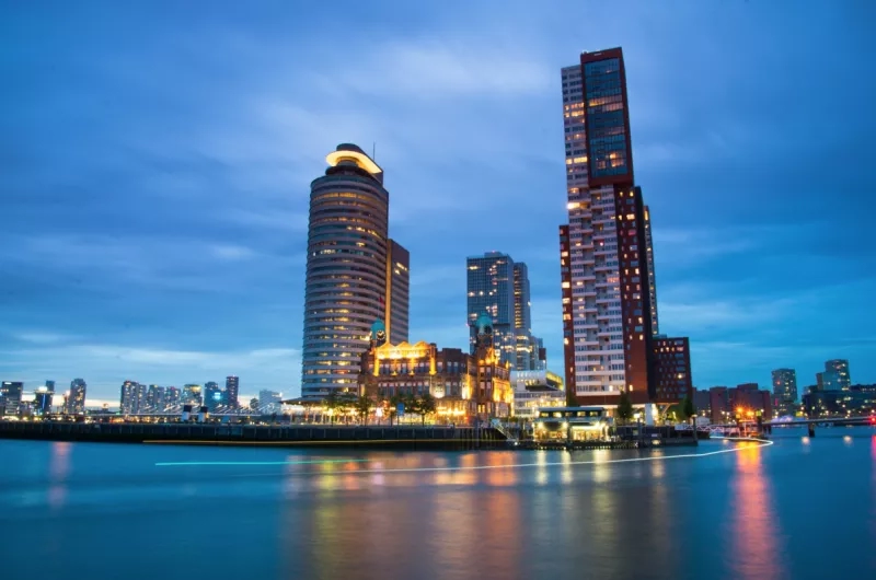 A cityscape at night with tall buildings, water, and a bridge, creating a vibrant urban ambiance.