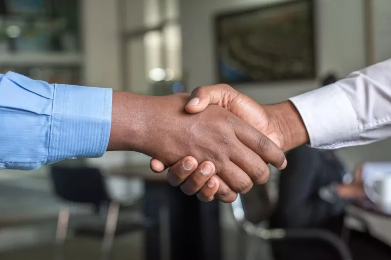 Two people shake hands in an office while another person sits nearby and a cup rests on a table.