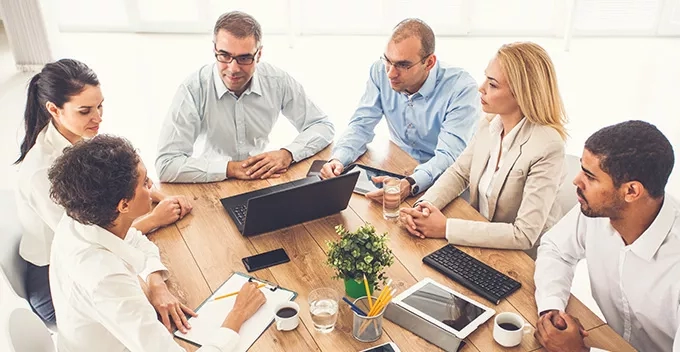 A group of people are sitting in a meeting room around a table with laptops and papers.