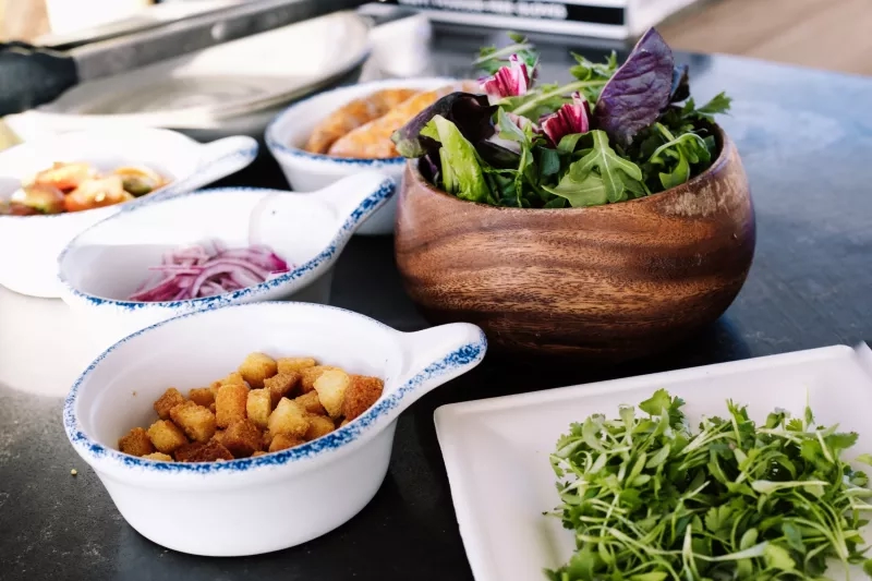 A wooden bowl of salad and four white bowls of salad toppings on a black table.
