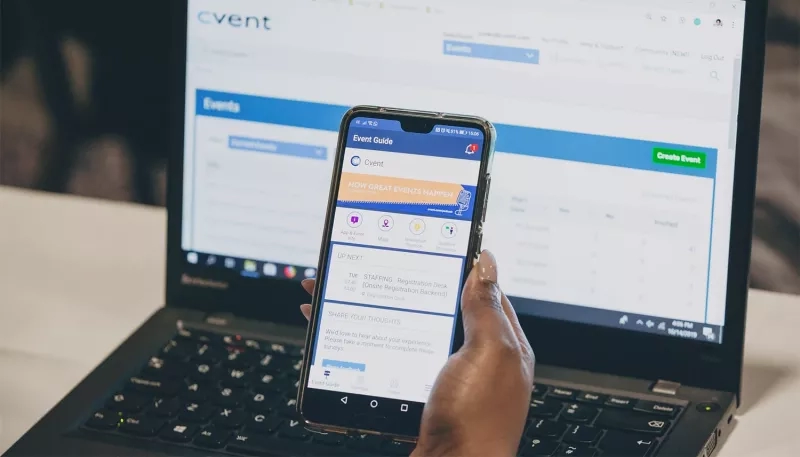 Hand holding a smartphone with the Event Guide app open in front of a laptop.