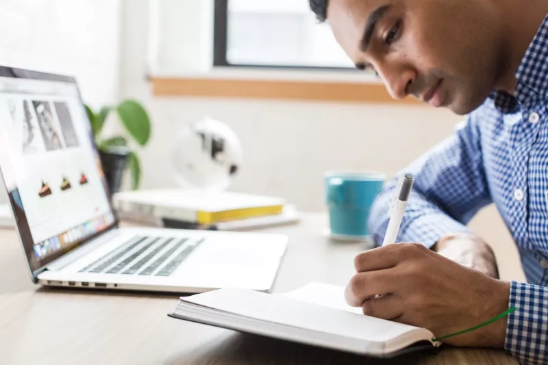 A man writing on a notepad while his laptop is open on the table next to him.