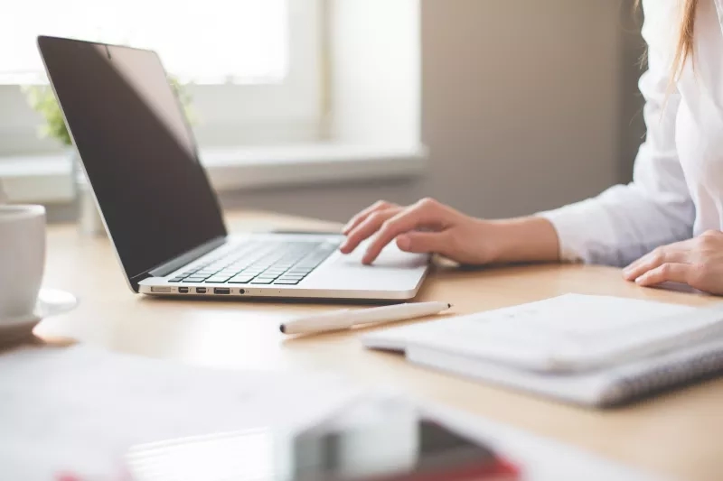 Woman working at desk with laptop, notebook, pen, and coffee cup, with natural light from window.