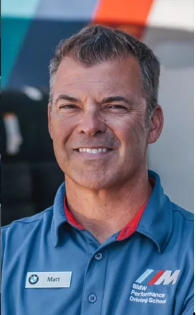 Smiling Matt wearing a blue BMW Performance Driving School polo shirt with a badge on the left side.