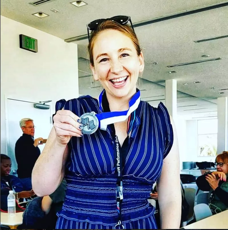 Woman holding a medal with a happy expression on her face in an indoor room with other people.