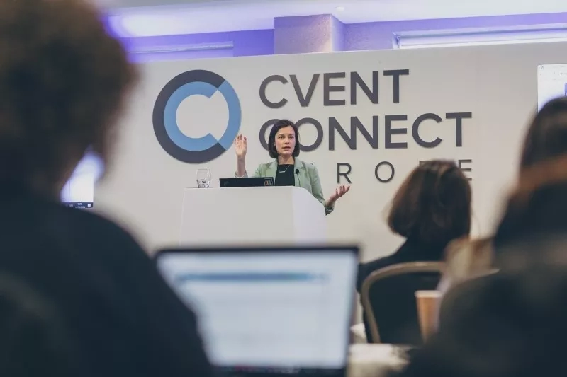 Woman giving a presentation in front of a Cvent Connect conference banner in a room.