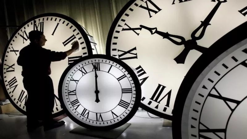 A man adjusts clocks in front of several large clocks with roman numerals and black hands.