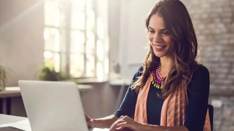 A woman sits at a desk, smiling, and typing on a laptop in a brick-walled room.