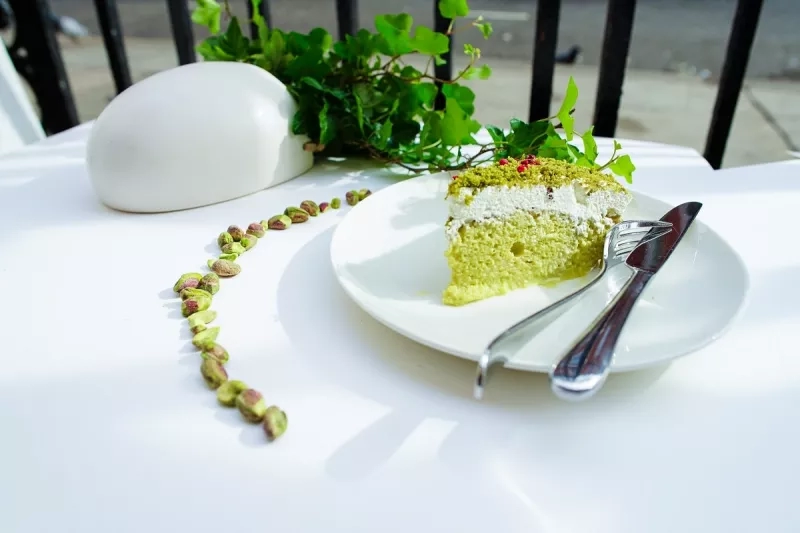 A slice of pistachio cake on a plate with a knife and fork on a white table.