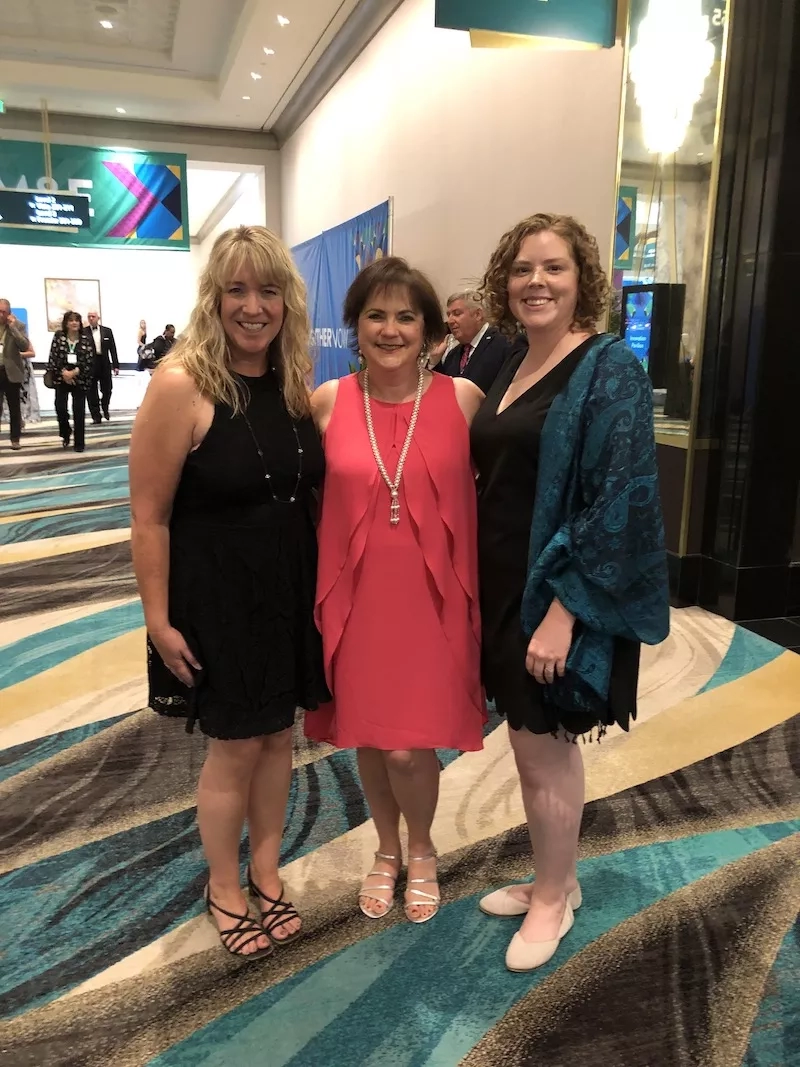 Three women stand together on a colorful carpet in front of a mirror in a lobby.