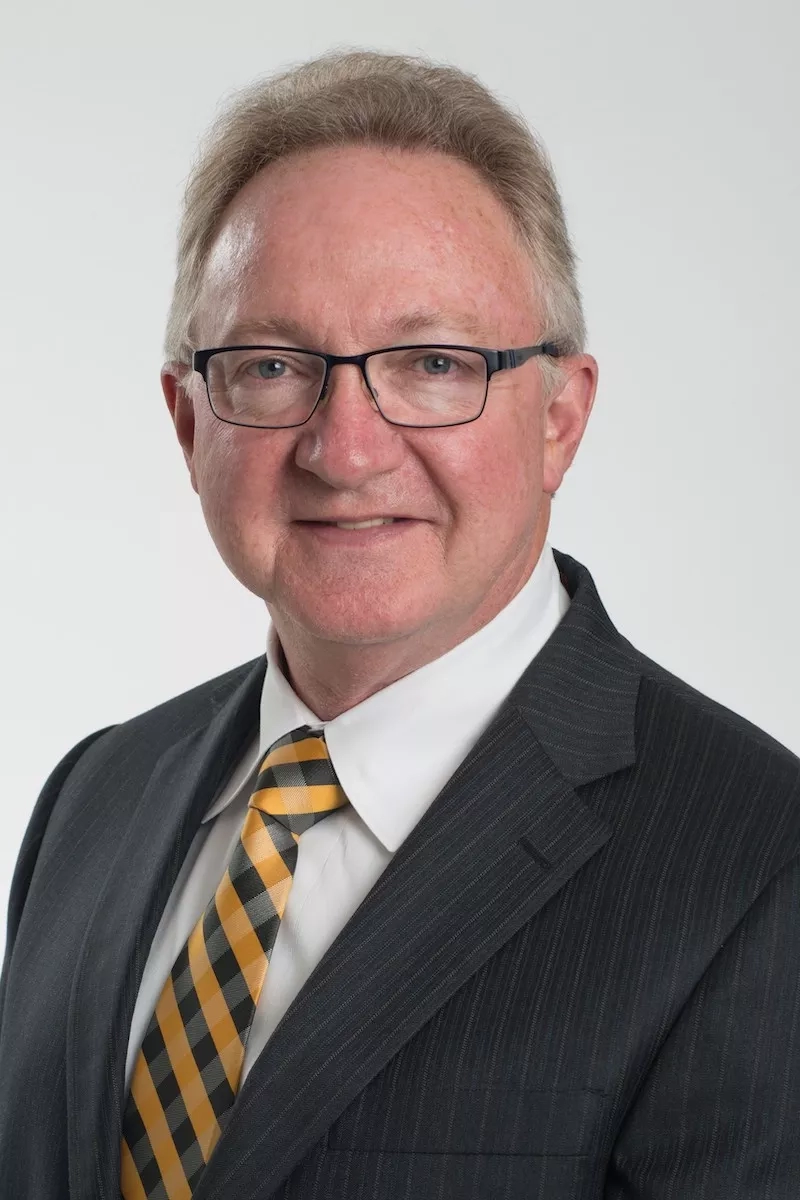 Kyle Flinn, a male, with short hair and glasses, wearing a suit, shirt, and yellow tie.