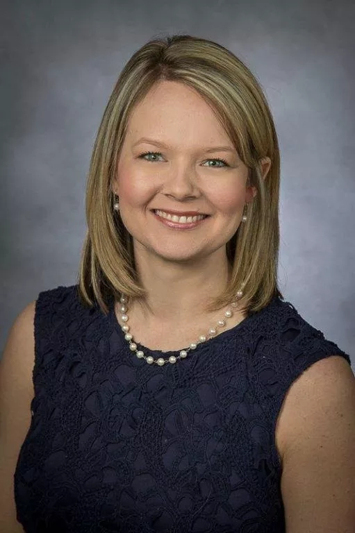 A headshot of a smiling woman in a navy blue dress with a pearl necklace and earrings.