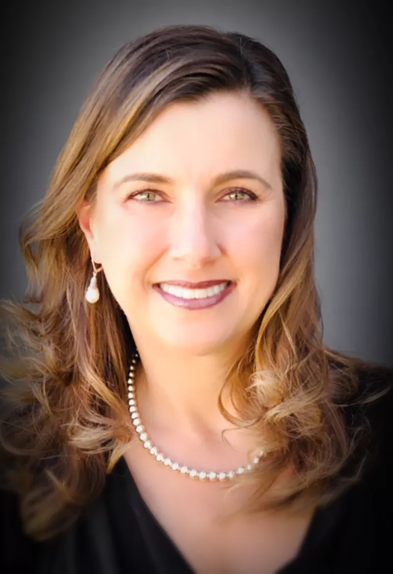 An image of a smiling woman wearing a pearl necklace and earrings on a black background.
