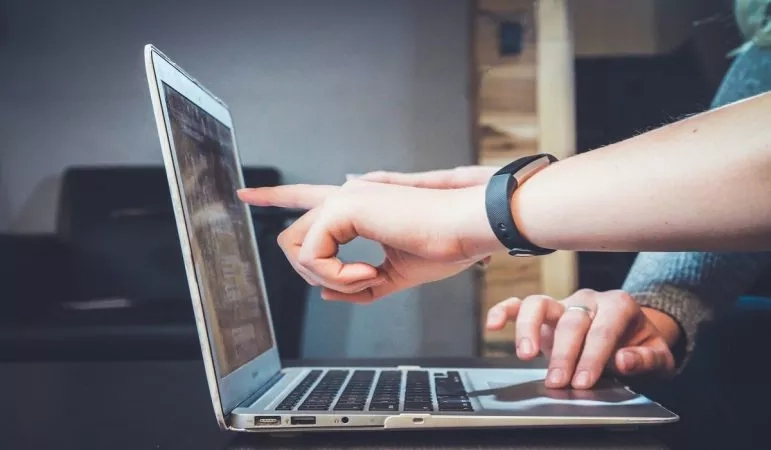 Two hands pointing to a laptop screen, one wearing a fitness tracker, in a room with wooden elements.