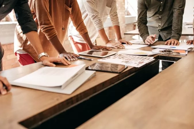 People working together in an office setting, with papers, pens, and tablets on a wooden table.