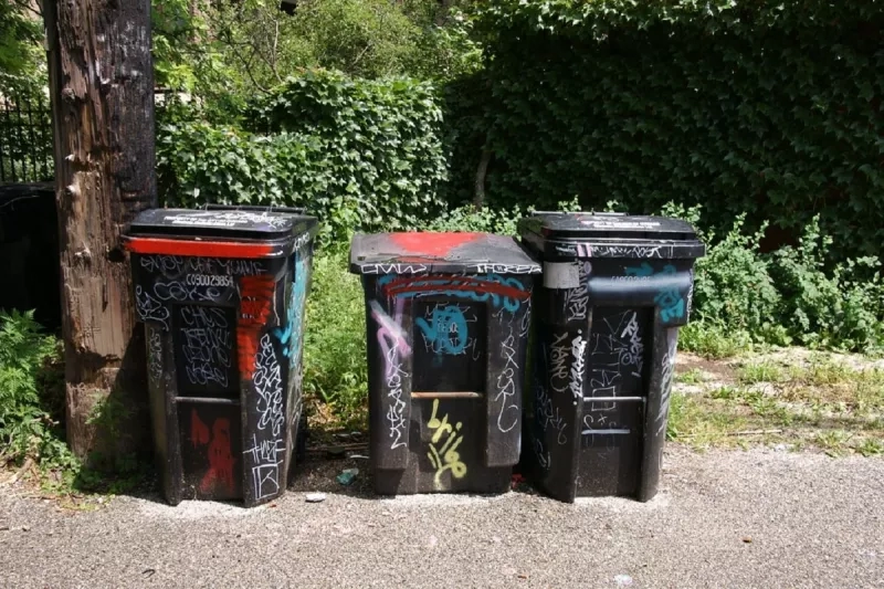 Three trash bins with graffiti on them are on the side of the road with trees in the background.