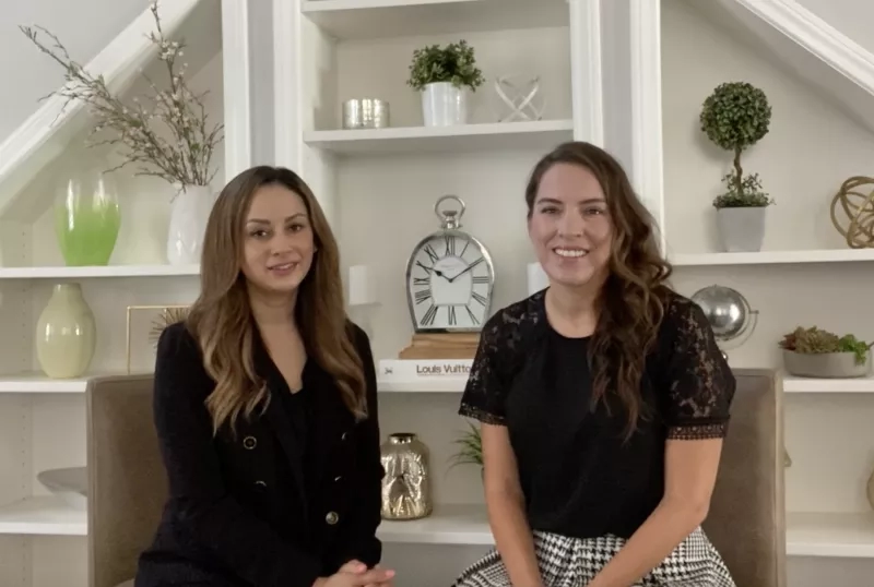 Two women sit on a couch in front of a shelf with vases, plants, and a clock.