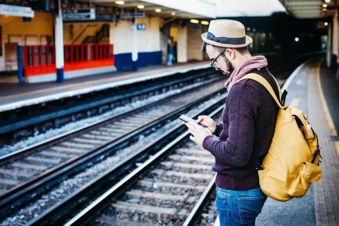 A man wearing a yellow backpack is standing at a train station and looking at his phone.