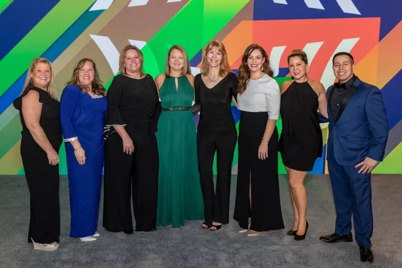 Seven smiling employees standing next to each other in front of a colorful screen at a company event.