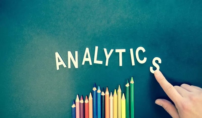 A hand points to the letter "s" of the word analytics, written above colored pencils.