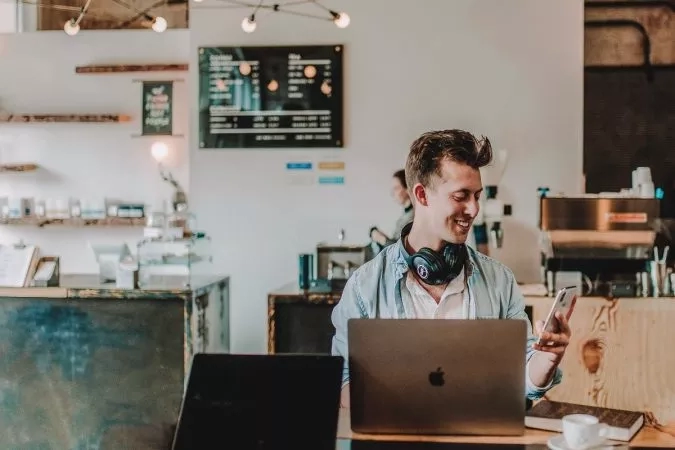 Man with headphones in a cafe, using a laptop and a smartphone, smiling, and having a cup of coffee.