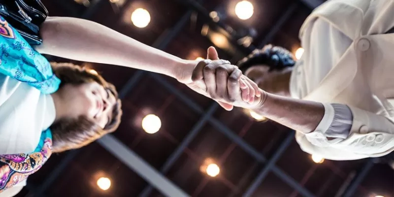 Two people hold hands as they dance in a dimly lit room with a geometric ceiling and yellow lights.