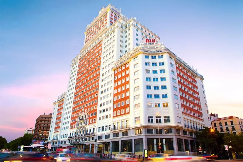A white and orange hotel building, called Riu Plaza Espana, in a city surrounded by trees and busy roads.