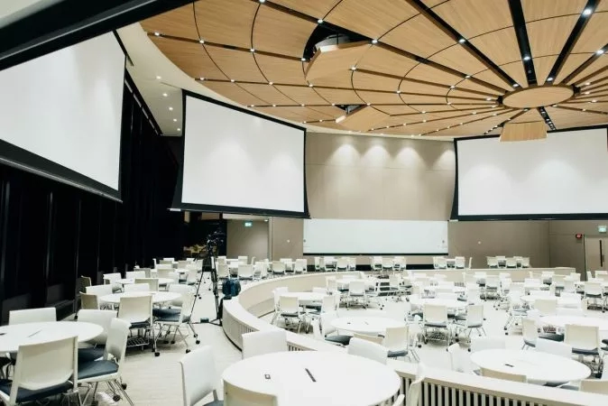 A conference room with tables and chairs, a tripod, and three projection screens on the walls.