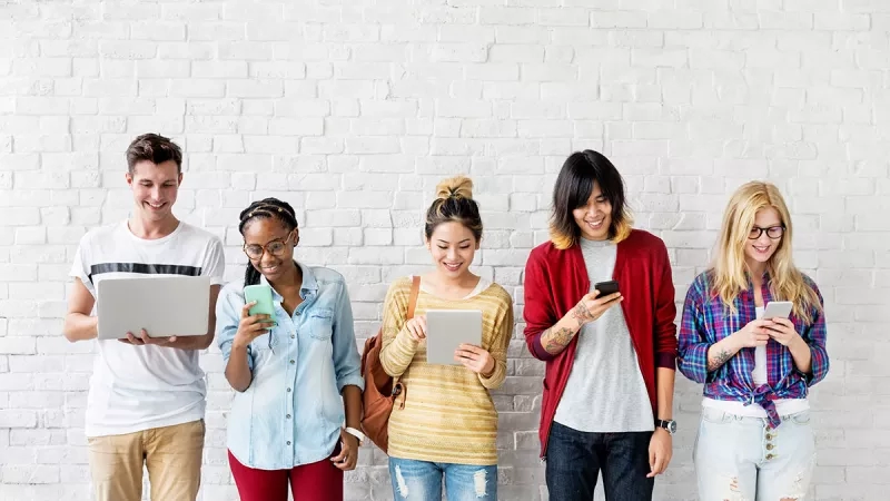 Five people standing in a row, each holding a tablet or smartphone, smiling and looking at their devices.